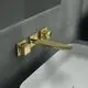 Wand-Waschtischarmatur in gebürstetem Gold von Ideal Standard mit eckigen Rosetten und einem langen, flachen Auslauf, montiert an einer grauen Wand über einem weißen Waschbecken.